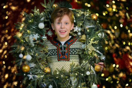 Happy smiling boy with a big decorated wreath around his neck posing on a festive Christmas tree and lights background. Merry Christmas and Happy New Year celebration.の写真素材