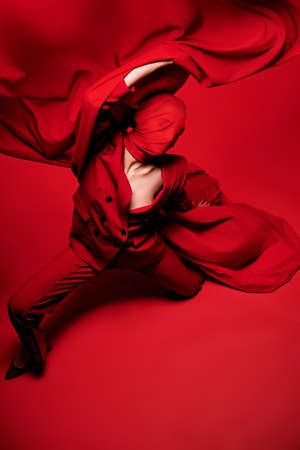 Fashion shot of a female high fashion model in pantsuit and red chiffon fabric covering her face expressively posing in motion on a red studio background.の写真素材