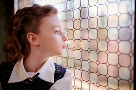 Portrait of an elegant girl child in classic school uniform looking through the window in a vintage room. Kid's fashion.の写真素材