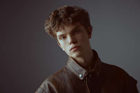 Men's youth fashion. Portrait of a handsome male model posing in a brown leather jacket on a grey background in the studio. Fashionable autumn clothes.の写真素材