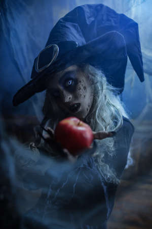 Halloween. A terrible wicked witch holds a poisoned apple in her hands, standing in her lair among the cobwebs. Evil black magic.の写真素材