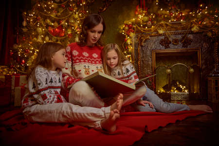 Happy Christmas family. A beautiful mother reads fairy tales to her beloved daughters, sitting at home by the fireplace and Christmas tree on Christmas Eve.の写真素材