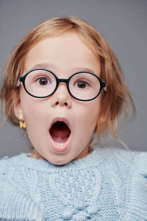 Portrait of a cute seven-year-old girl with fair hair and stylish glasses with a big surprise on her face posing on a gray background. Education, Junior School. Glasses style.の写真素材