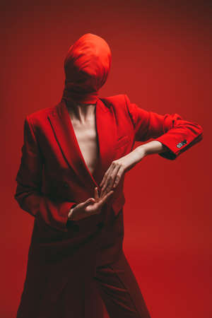 Graceful fashion model girl posing in a smart red pantsuit with her face hidden behind red chiffon cloth on a red studio background. High fashion and art.の写真素材