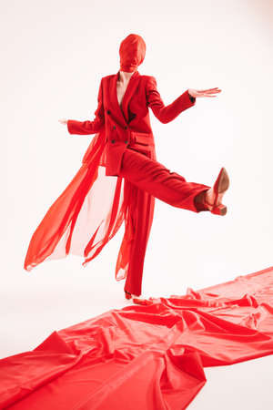 Stunning female fashion model in a smart red pantsuit and cloth hiding her face expressively walks a red carpet. Full length shot on a white background. Haute couture fashion.の写真素材