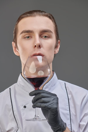 Professional sophisticated male chef in neat uniform carefully examines red wine in a glass.