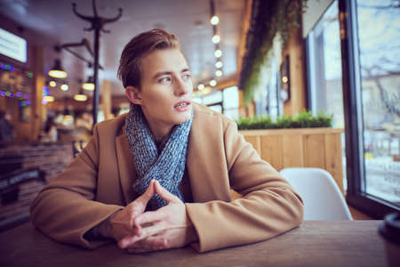 Portrait of a handsome blond man  in stylish winter clothes sitting at a table in a cozy cafeteria on a cold winter day and looking out the window. People concept, lifestyle.の写真素材