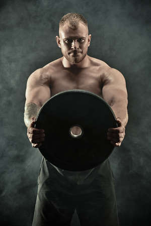 Sports concept: weightlifting, bodybuilding. Portrait of a muscular handsome athletic man holding a large iron disk in his hands. Black background with smoke.の写真素材