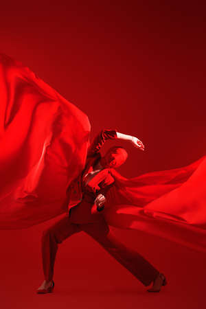 Expressive fashion model with her face wrapped in red cloth posing in red suit and fluttering chiffon fabric. Full length studio portrait on a red background. High fashion and art.の写真素材