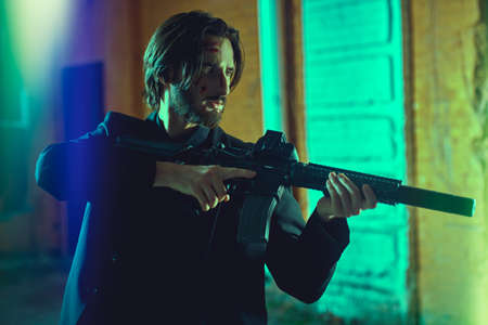The criminal world of the big city. Courageous man with wounds on his face chases criminals with a rifle in his hands in an industrial zone at nighttime. Special agent.の写真素材
