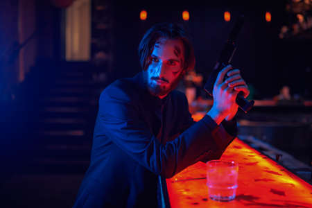 Portrait of a handsome wounded special agent drinking whiskey in a bar and holding his gun. Action movie hero.の写真素材