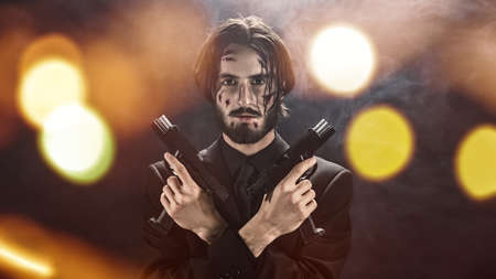 Handsome wounded special agent in a black suit holds guns in his hands in a dark smoky background with flickering lights.の写真素材