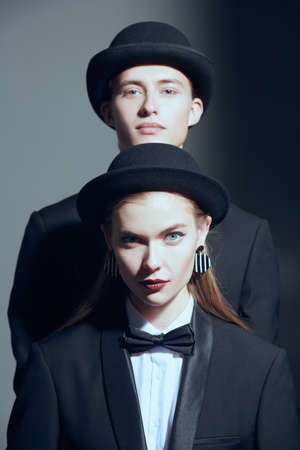 Perfect people look. Fashion models man and woman posing in elegant black pantsuits, white shirts, bow ties and black bowlers on a grey background. Men's clothing style. Fashion shoot.の写真素材