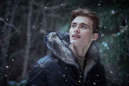 Portrait of a young blond man in a trendy black leather jacket with a fur hood standing in the park and admiring the beauty of the nature on a snowy winter day. Men's fashion. Winter season.の写真素材