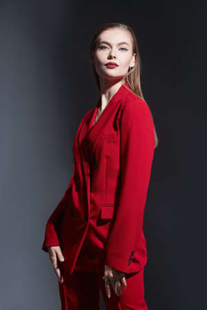 Elegant business style. Portrait of a beautiful young woman with elegant makeup posing in smart red pantsuit on a dark gray background. Femininity, feminism and fashion.の写真素材