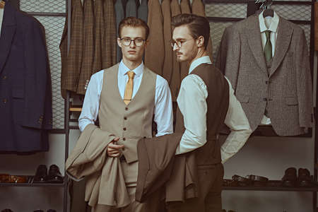 Men's fashion. Two well-groomed successful businessmen in classic suits and glasses stand in a premium men's clothing store.の写真素材