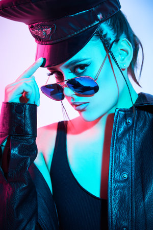 Stunning brunette woman with trendy sunglasses poses in a black leather jacket and peaked cap on a...