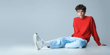 Attractive male model poses in a red sweater, light blue jeans and and sneakers sitting on the floor in the studio. Men's youth fashion. Copy space.の写真素材
