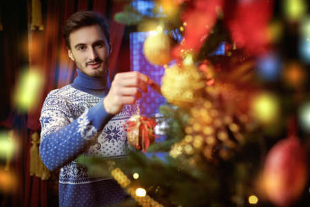 Good-looking smiling young man in warm winter sweater joyfully decorates a Christmas tree. Merry Christmas and Happy New Year!の写真素材