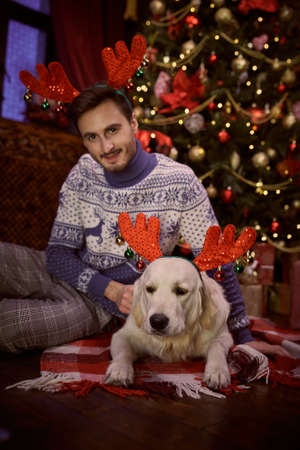 Happy owner and his pet. Portrait of a joyful young man and his pet dog in funny xmas deer horns sitting in a beautiful cozy room by the Christmas tree.の写真素材