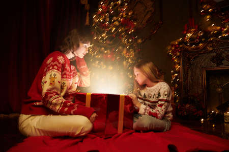 Believe in magic on Christmas Eve. Happy mom and little daughter sit by the fireplace and the Christmas tree looking with interest into a gift box with magic light coming out of it.の写真素材