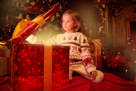 Magic Christmas time. Cute little girl opens the cover of a gift box with magic light coming out of...