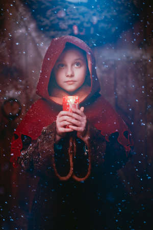 Believe in a miracle at Christmas. An angelic little girl in medieval clothes stands under the snow at a wooden house on Christmas Eve with hope in her eyes. A beam of light falls on her face.の写真素材