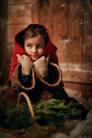 Believe in a miracle at Christmas. An angelic little girl in medieval clothes sits at a wooden house on Christmas Eve with fir-tree branches in her basket. Magic Christmas night with heavy snowfall.の写真素材