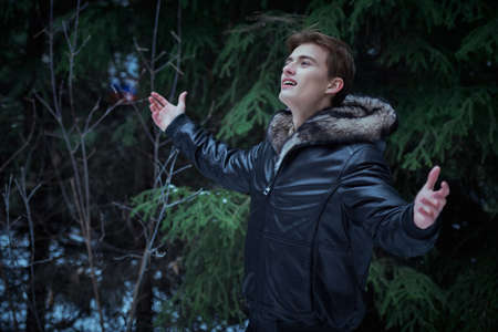 Young blond man in a trendy black leather jacket with a fur hood stands with outstreched arms in the winter park admiring the beauty of the nature. Men's fashion. Wonderful winter season.の写真素材