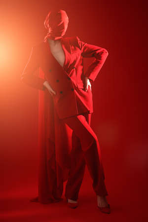 High fashion female model elegantly posing in red pantsuit and red chiffon fabric hiding her face. Full length shot with bright light shining on a red background. Fashion and red color.の写真素材