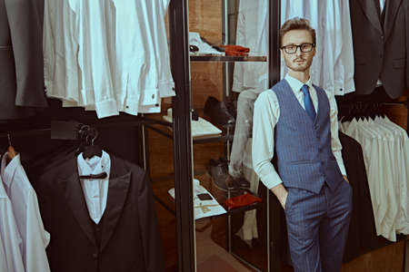 A handsome brunet man in elegant classic suit and spectacles stands relaxed in a premium men's clothing store. Men's business fashion.の写真素材