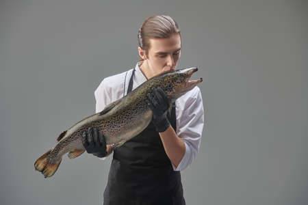Food and meals. Young male chef in a neat uniform and apron kisses a big fresh salmon in his hands. Grey studio background.の写真素材