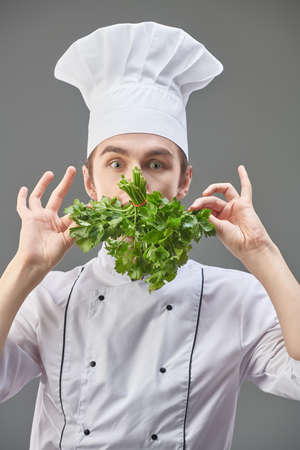 Artistic young chef in neat uniform holds a bunch of parsley next to his face hiding his mouth. Studio portrait on a grey background.の写真素材