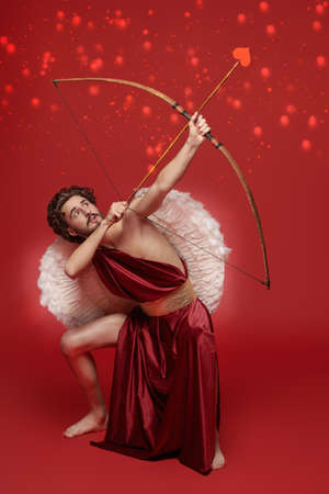 Valentine's Day concept. Full length portrait of the God of love - Cupid aiming up with bow and arrow on a red background.の写真素材