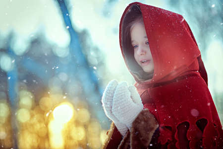 Winter's tale. A cute girl in warm medieval clothes with a cape and a hood stands frozen in a snowy forest warming her hands. The sun is setting in the background.の写真素材