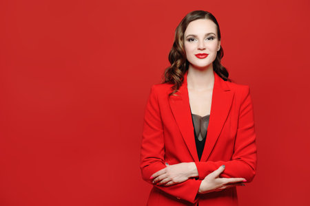 Elegant business style. Portrait of a beautiful confident young woman with bright makeup posing in smart red jacket on a red background. Femininity, feminism and fashion. Copy space.の写真素材