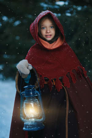 Christmas Fairy Tale. Cute medieval girl in winter clothes with a cape and a hood walks through a snowy spruce forest with a burning lantern lighting her way. Historical reconstruction.の写真素材
