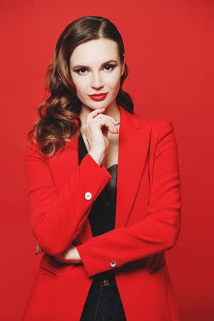 Beautiful confident woman with elegant makeup and hairstyle posing in bright red classic blazer on a red background. Femininity, feminism and fashion. Elegant business style.の写真素材