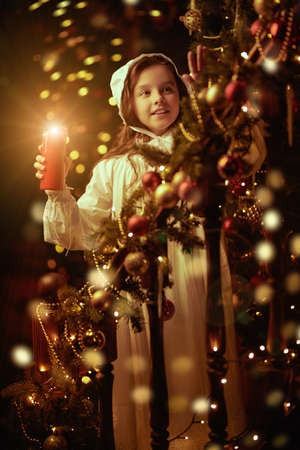 Joyful little girl in a nightie and nightcap stands on the staircase with a burning candle looking at the beautiful Christmas decorations in a vintage room. Vintage style. Christmas and New Year concept.の写真素材