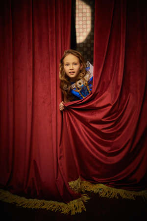 A cute joyful girl with beautiful golden curly hair in a princess dress excitedly looks out from behind velvet curtains. Vintage room. Christmas tale.の写真素材
