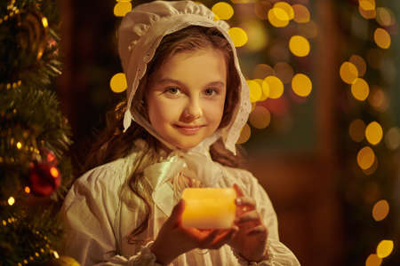 Winter Christmas Tale. Portrait of a joyful little girl in a nightie and a nightcap, standing with a candle in her hands in a fabulous Christmas atmosphere and smiling sweetly. Portrait in vintage style.の写真素材