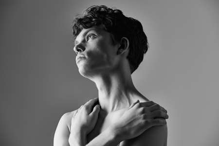 Black and white photo of a young man with wavy hair crossing his arms on his bare chest. Male beauty and health.の写真素材