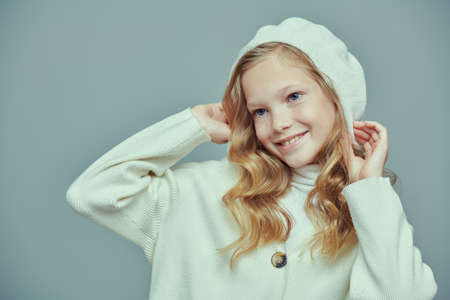 Cute joyful 11 year old girl with blonde hair and freckles poses in a studio in a stylish white beret and sweater. Fashion shot on a gray background. Kid's fashion.の写真素材