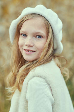Autumn. Portrait of a cute smiling little girl with wavy blonde hair dressed in a stylish warm hat and classical clothes posing in an autumn park. Children's fashion.の写真素材