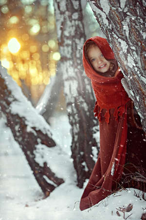 A cheerful medieval girl in caftan and cape joyfully peeks out from behind a tree in a snowy forest at the sunset. Fairy tales on Christmas and New Year.の写真素材