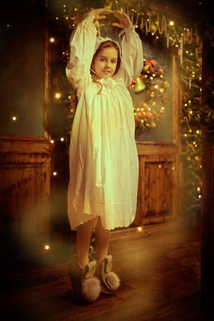 Once upon a time at Christmas. A happy little girl in a nightie and a nightcap is dancing in a fabulous Christmas setting and smiling. Full length portrait in vintage style.の写真素材