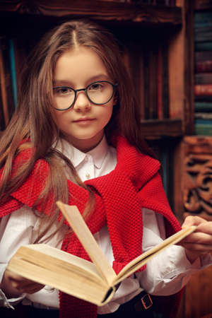 Pretty little girl in elegant classic school uniform and glasses poses in a luxurious vintage library interior a with book in her hands. Kid's school fashion.の写真素材