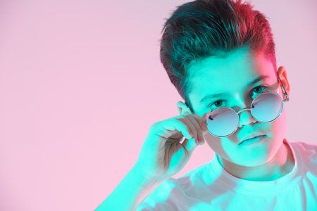 Portrait of a handsome boy teenager in white t-shirt and stylish sunglasses looking at camera seriously. Studio portrait in green and pink light. Copy space.の写真素材