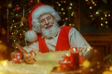 Christmas tale. Kind old Santa Claus answers children's letters writing in an old scroll at his festively decorated cozy home surrounded by magic flickering lights.の写真素材