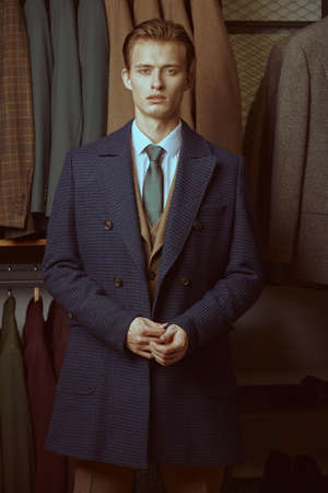 Fashion shot of a serious handsome blond man in elegant coat and three-piece suit standing in the background of luxury classic suits in a store. Men's fashion and style.の写真素材